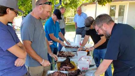 Pastor Jonathan Spronk, lower right in dark shirt and apron, leans in and places meat on a plate.