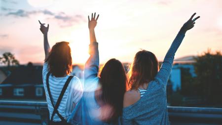 Three girls stand arm in arm with their backs to the camera. One is holding up a peace sign with her hands.