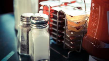 A salt and pepper shaker sit on a table in a diner beside jam and a ketchup bottle
