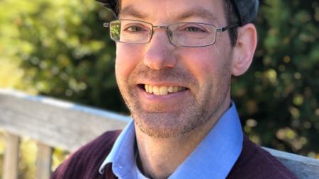 headshot of Mike, a white mane wearing glasses, a cap, and blue collared shirt with burgundy sweater 