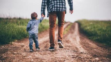 a child holds the hand of a parent while they walk down a dirt path in a field of grass