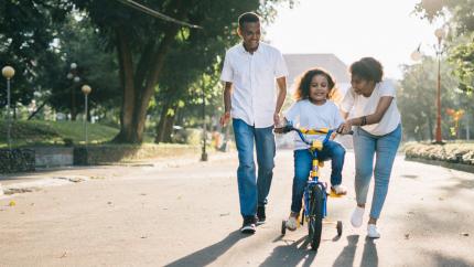 a mother and father help their child ride a bike on a sunny street