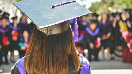 a woman with brown hair wearing a graduation cap and robe speaks to a group of graduates