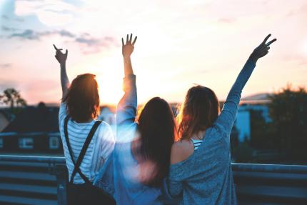 Three girls stand arm in arm with their backs to the camera. One is holding up a peace sign with her hands.