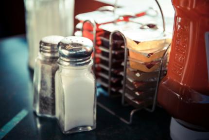 A salt and pepper shaker sit on a table in a diner beside jam and a ketchup bottle
