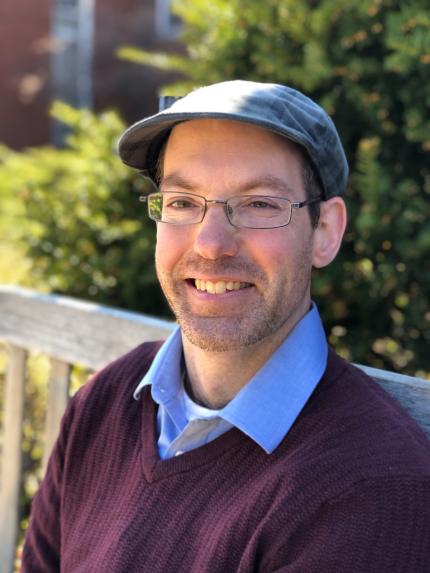 headshot of Mike, a white mane wearing glasses, a cap, and blue collared shirt with burgundy sweater 