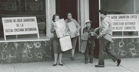black and white photo of a woman with brown grocery bags in front of a window with a sign reading "The Good Samaritan Center"